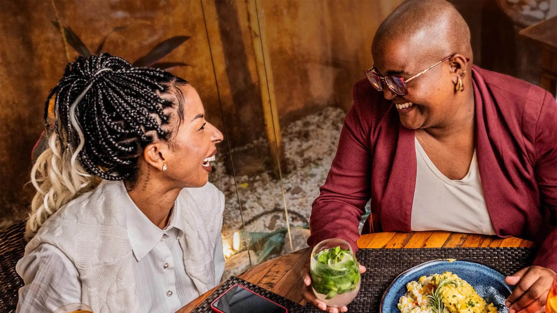 Two guests laughing together at a restaurant table with a cocktail and a plated dish